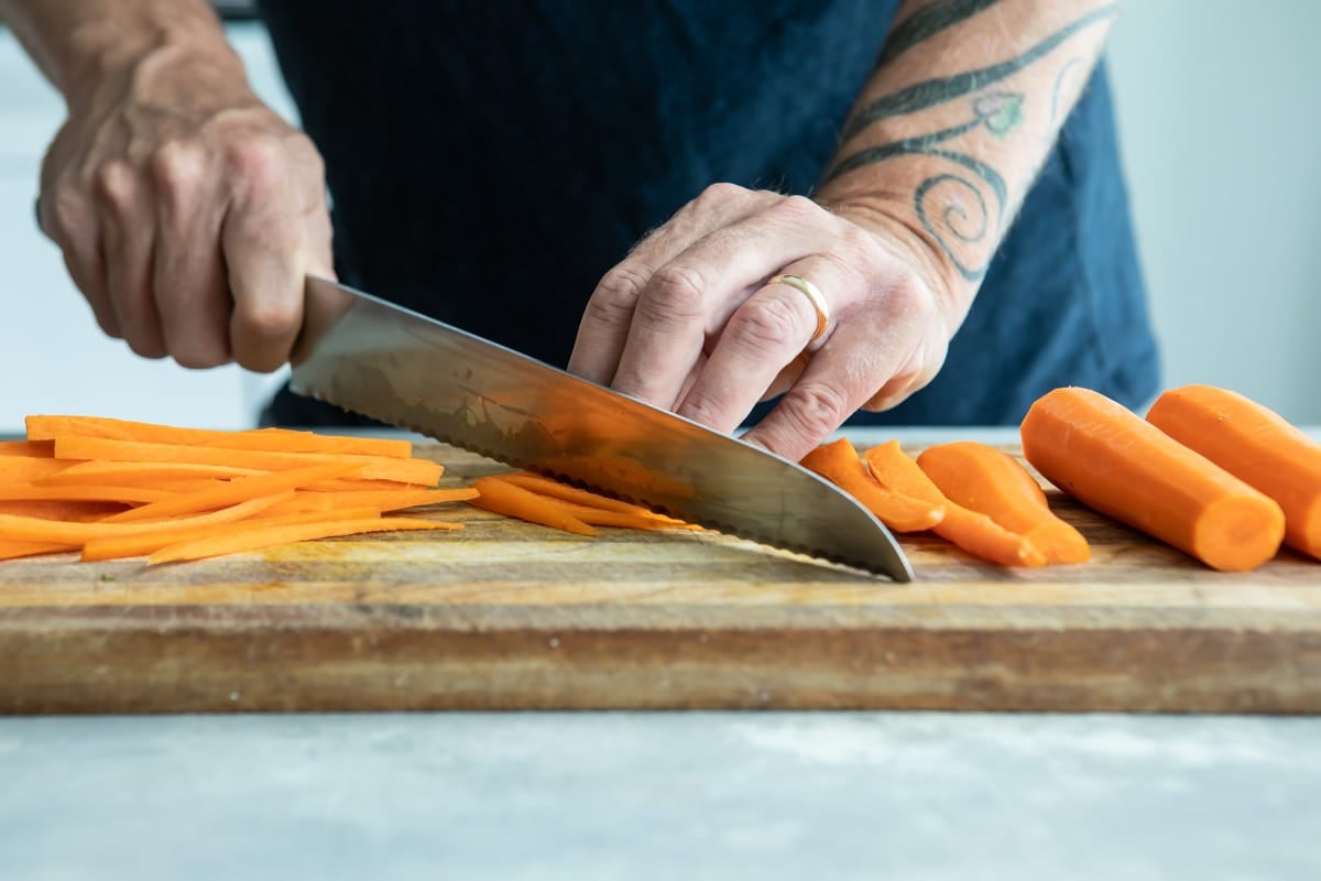 Someone cutting carrots on a wooden cutting board.