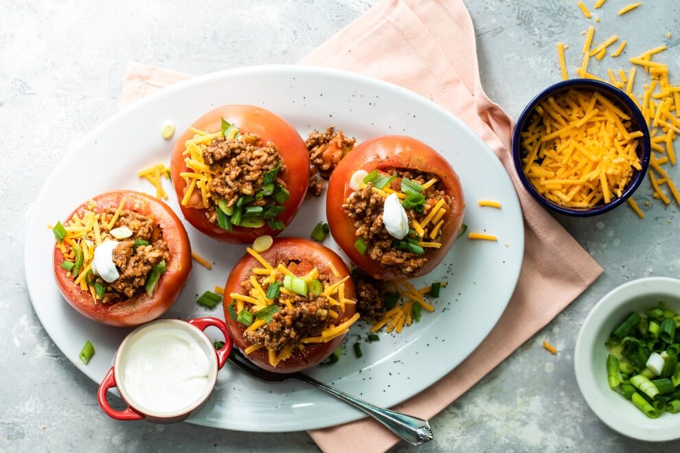 Four taco stuffed tomatoes on a white platter with a side of cheese, green onions, and sour cream.