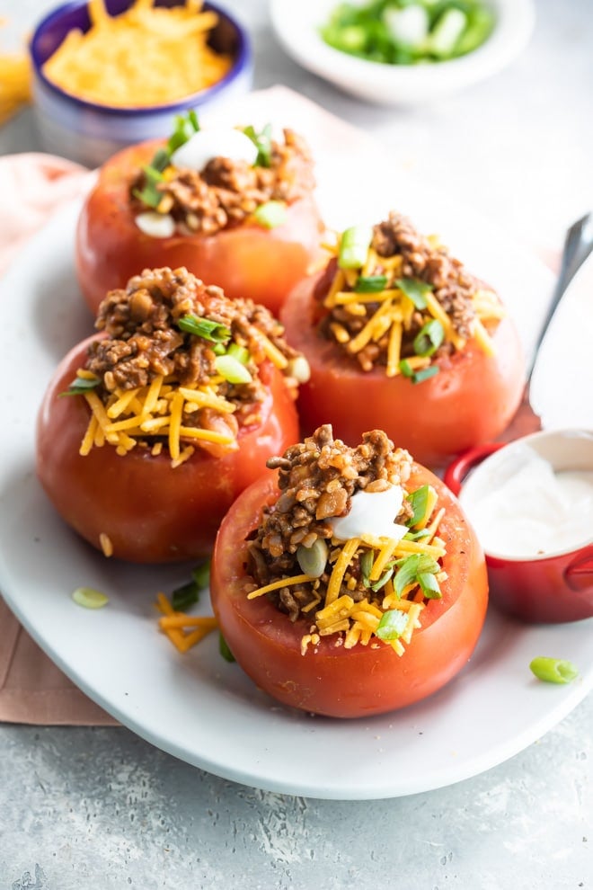 Four taco stuffed tomatoes on a white platter with a side of cheese, green onions, and sour cream.