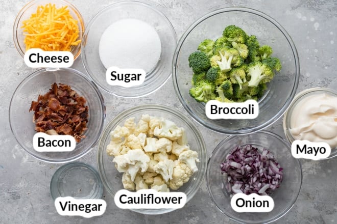 Ingredients for broccoli salad with bacon and cheese in various clear bowls on a gray counter.