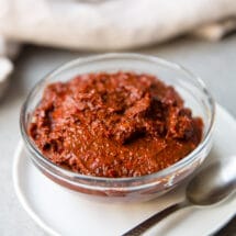 A bowl of harissa paste on a plate with a spoon next to it.