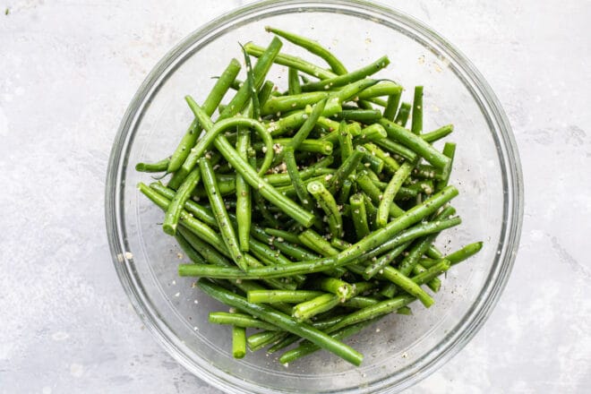 Green beans in a clear bowl coated with oil and seasonings.