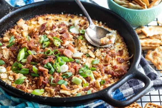 Cheesy bacon dip in a cast iron skillet surrounded by pretzels and crackers.