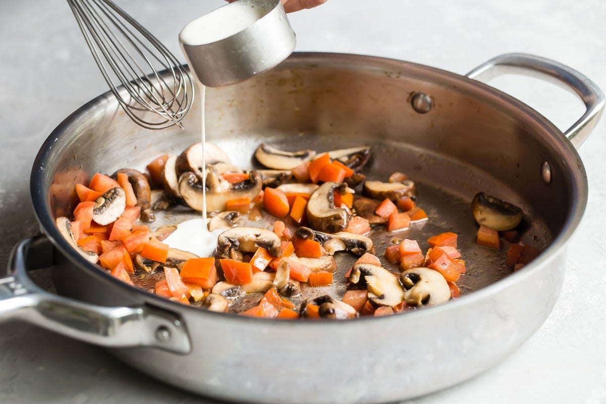 Adding cream to a skillet with cooked mushrooms and tomatoes.