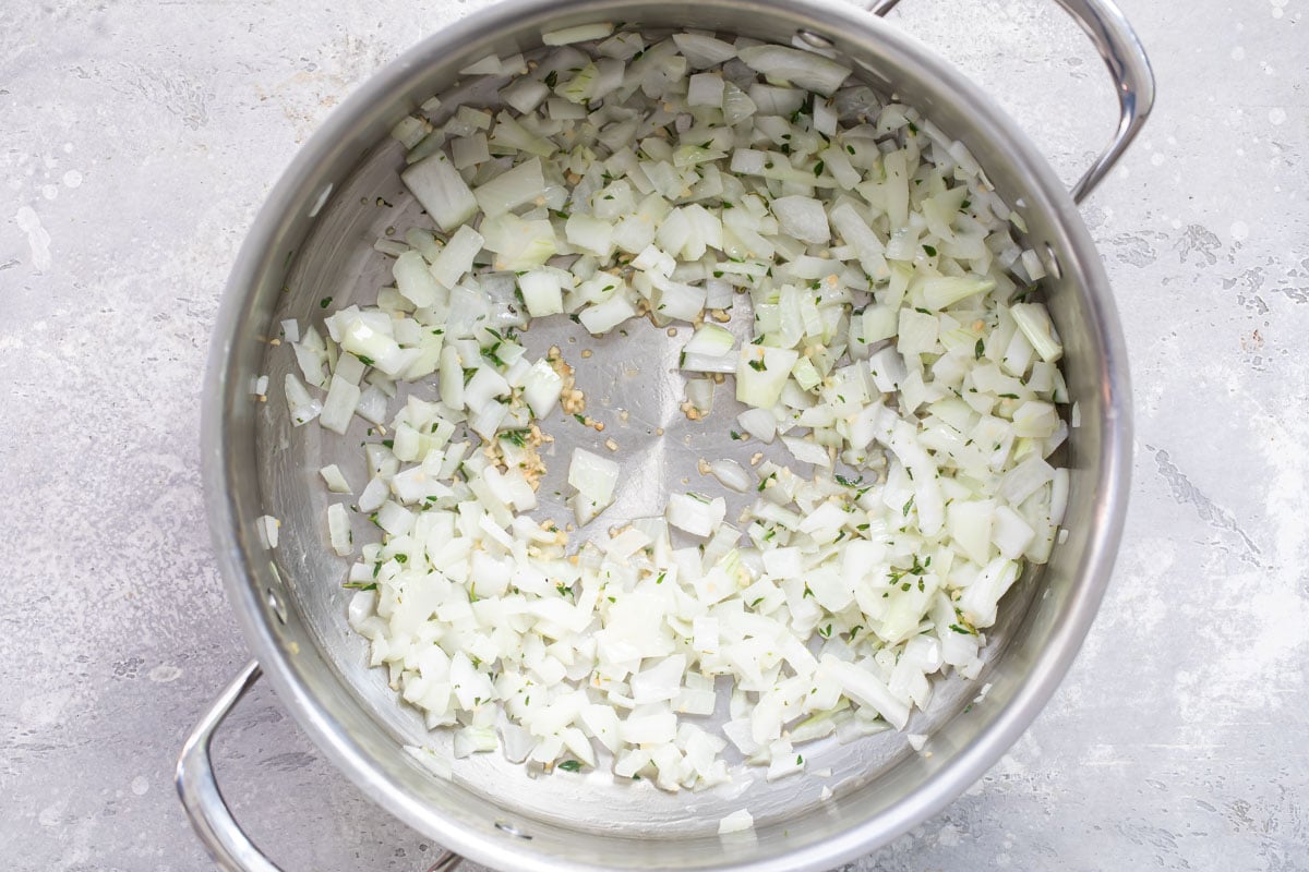 Onions, garlic and herbs being cooked in a silver stock pot.