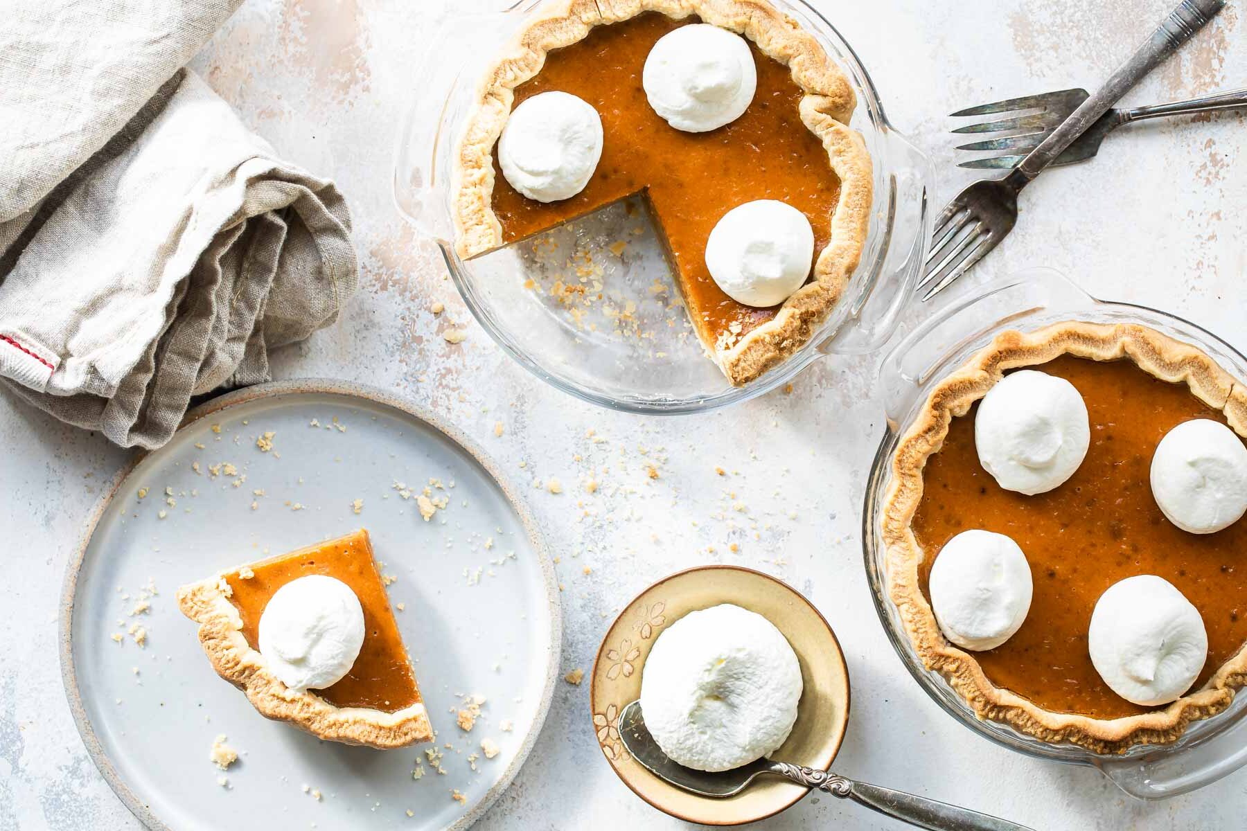 A slice of mini pumpkin pie on a white plate with two mini pumpkin pie and whipped cream behind it.