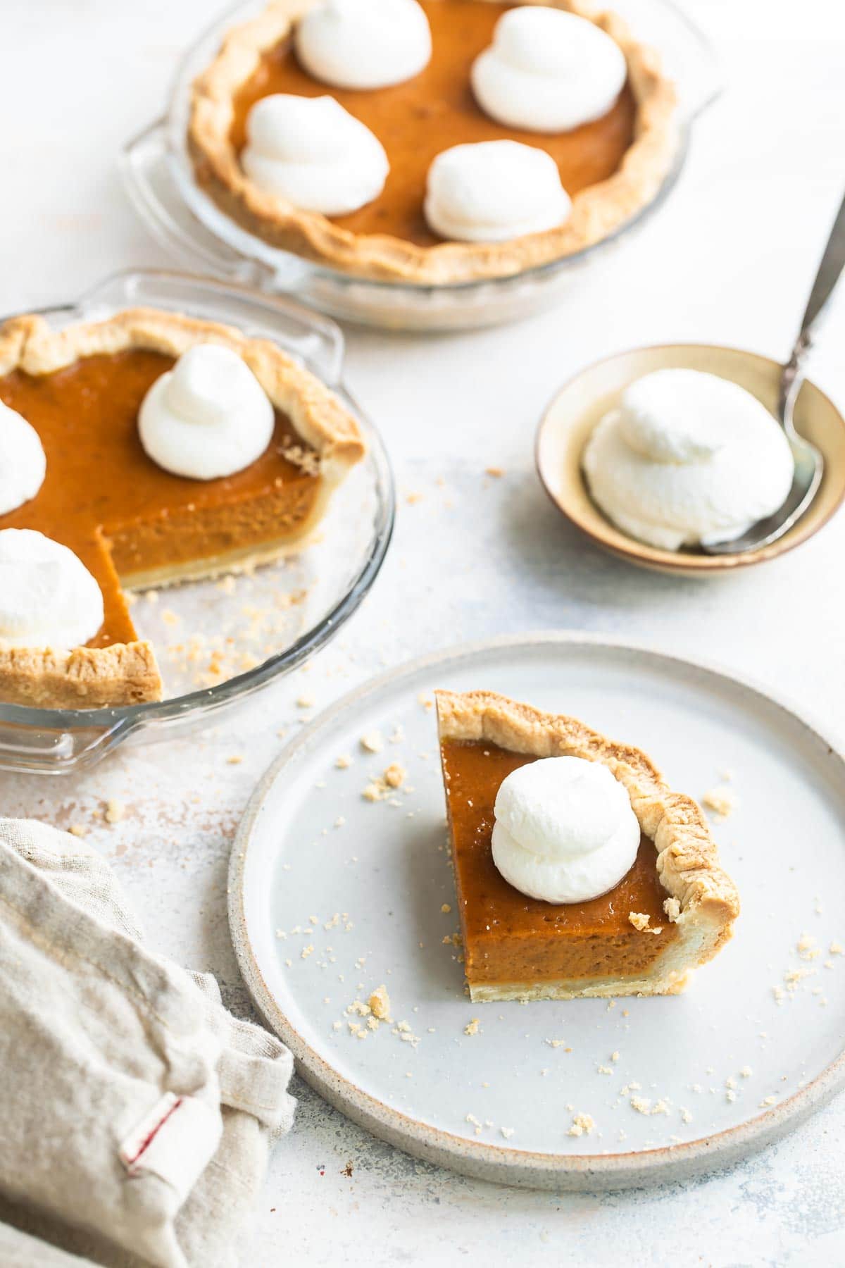 A slice of mini pumpkin pie on a white plate with two mini pumpkin pie and whipped cream behind it.