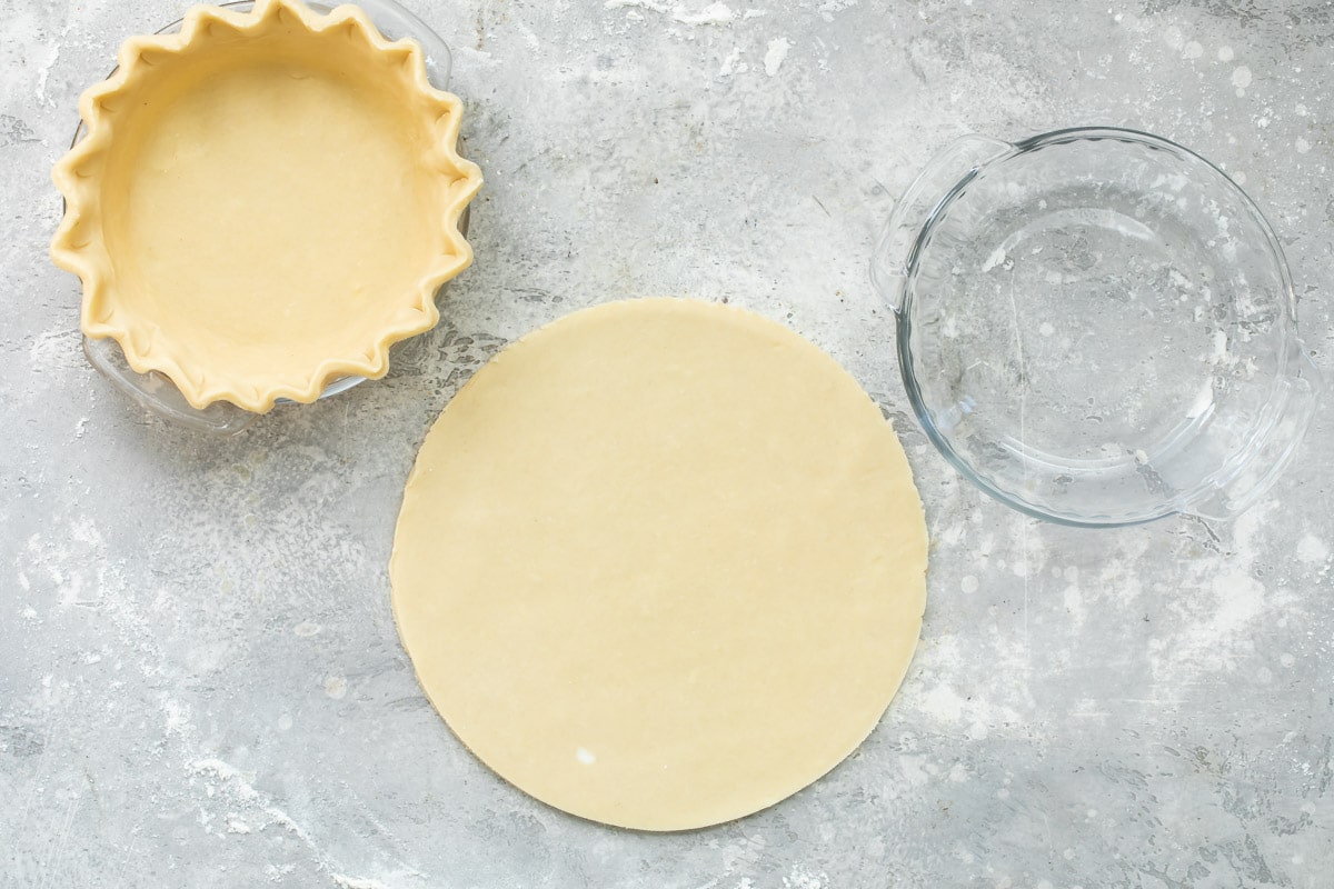 A mini pumpkin pie crust in a pie pan, a circle of pie dough, and an empty glass mini pie pan.