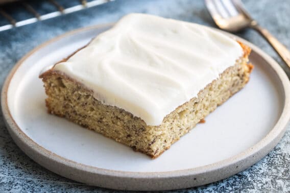 A slice of banana cake with cream cheese frosting on a white plate with a fork next to it.