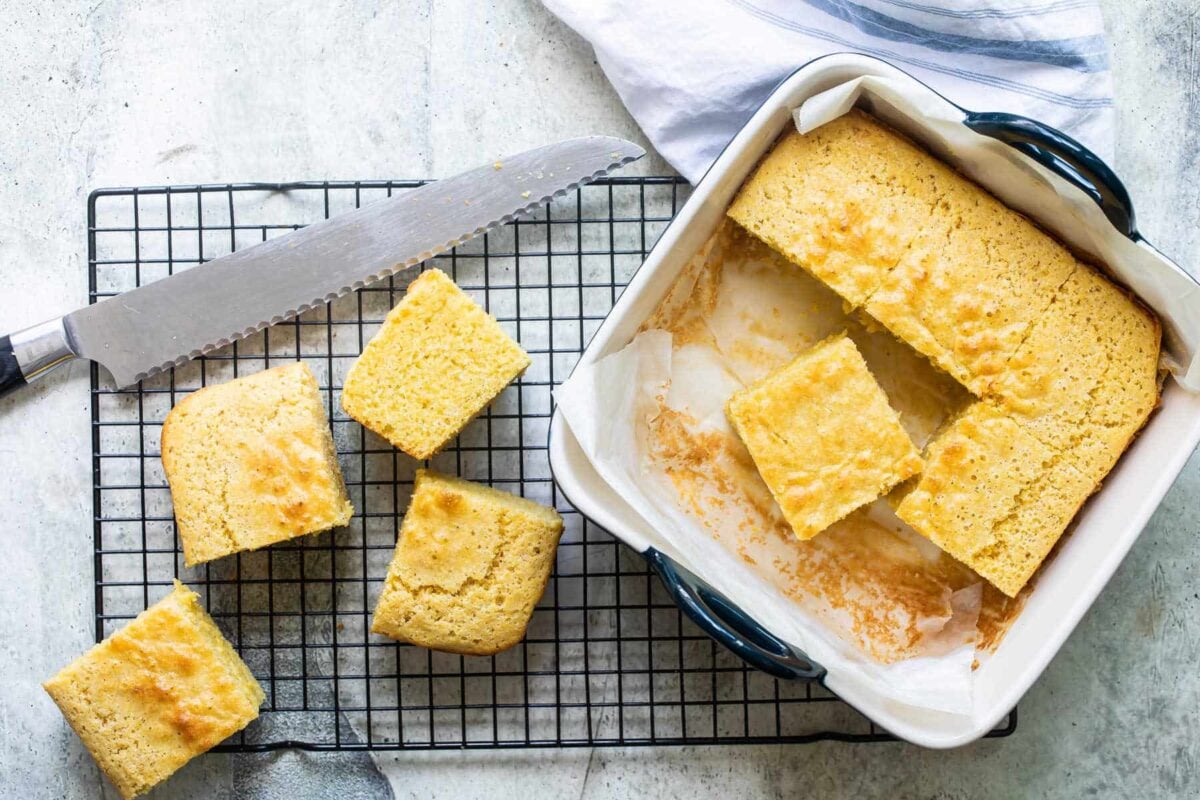 A pan of cornbread with four slices resting on a cooling rack with a bread knife.