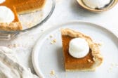 A slice of mini pumpkin pie on a white plate with a mini pumpkin pie and whipped cream behind it.