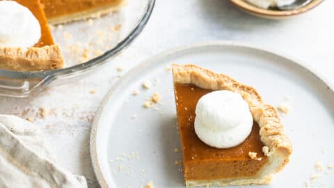 A slice of mini pumpkin pie on a white plate with a mini pumpkin pie and whipped cream behind it.