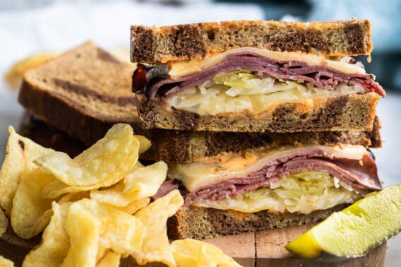 A reuben sandwich on a cutting board surrounded by chips and a pickle spear.