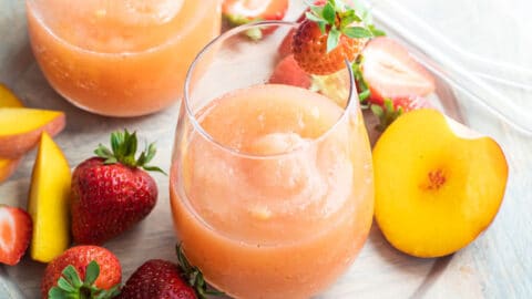 Two glasses of strawberry peach frose on a plate surrounded by slices of strawberries and peaches.