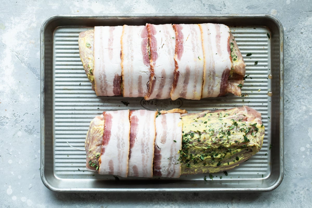 Two pork tenderloin on a baking sheet getting wrapped in bacon.
