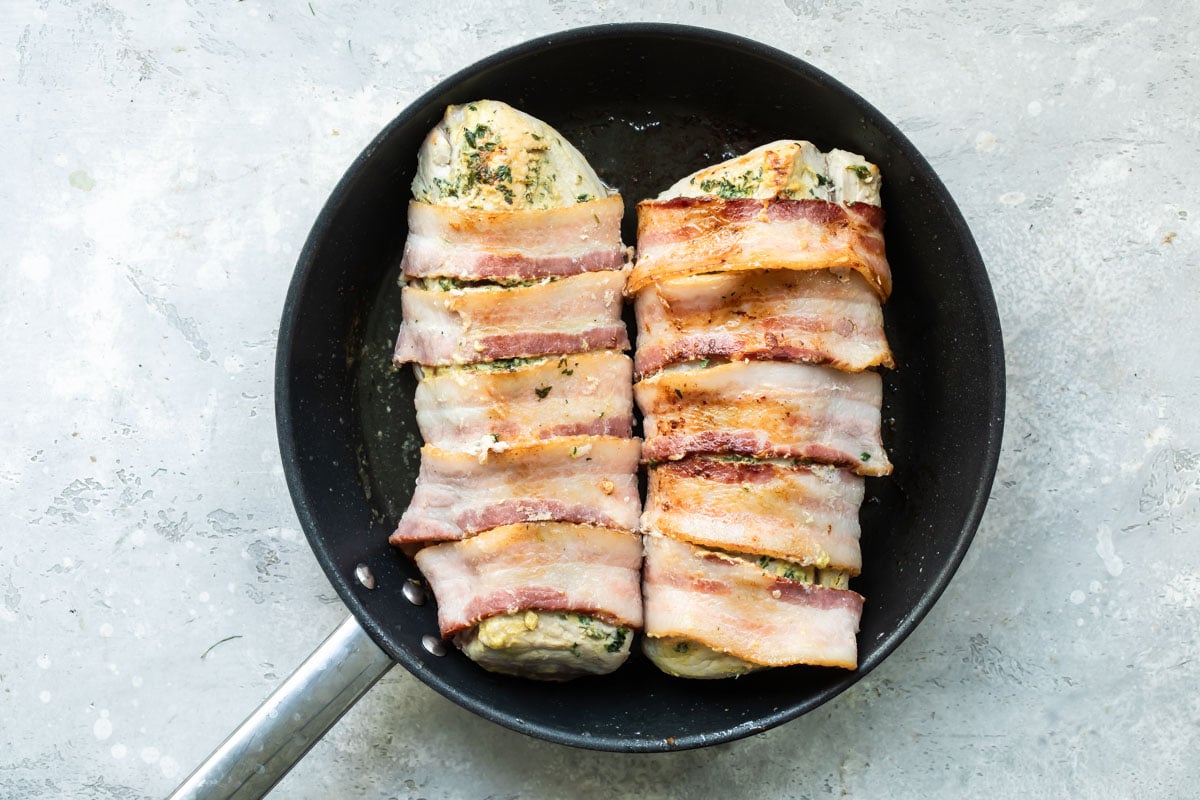 Uncooked bacon wrapped pork tenderloin getting seared in a black pan.