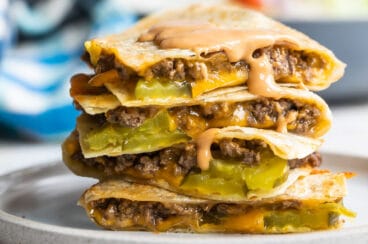 A cheeseburger quesadilla stacked on a plate with a wedge salad in the background.