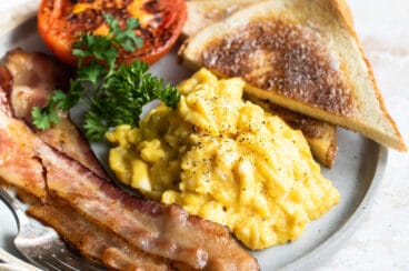 Scrambled eggs on a plate with toast triangles, bacon slices, parsley, and a roasted tomato.