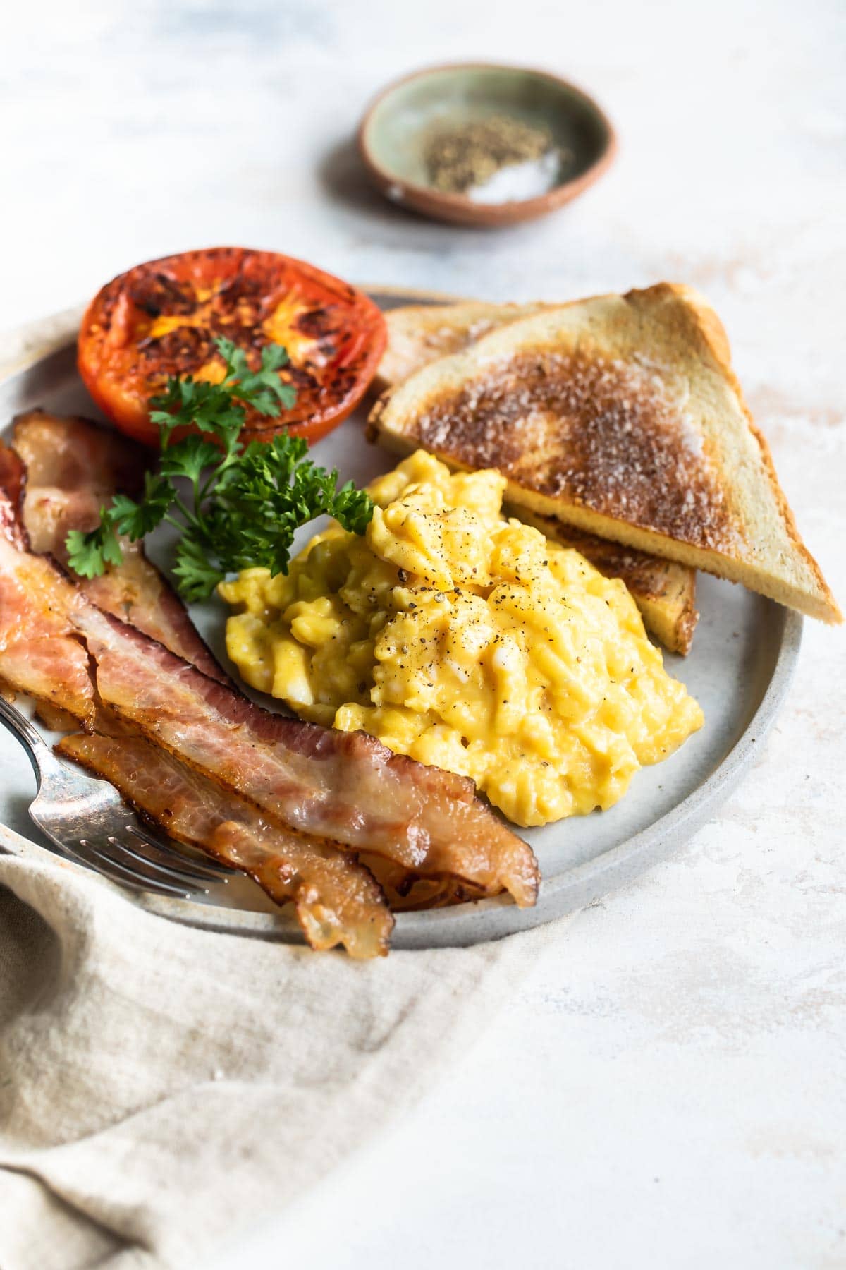 Scrambled eggs on a plate with toast triangles, bacon slices, parsley, and a roasted tomato.