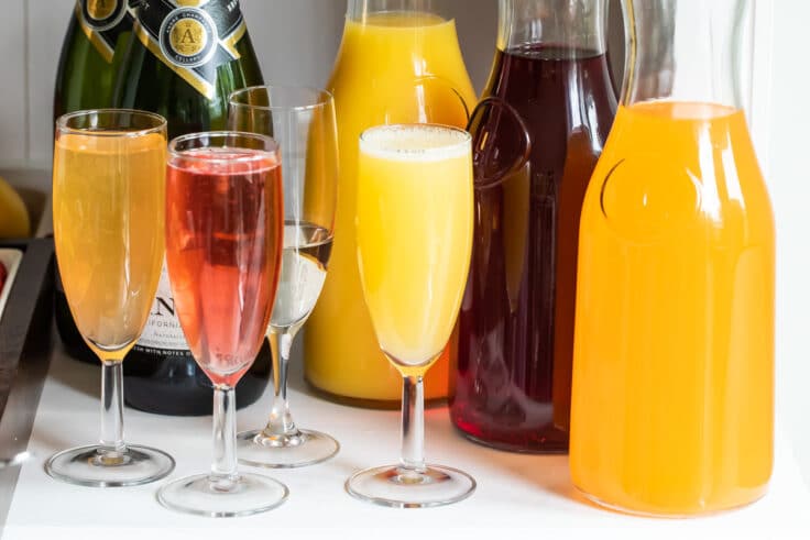 Three carafes with various juices and 4 champagne flutes with mimosas.