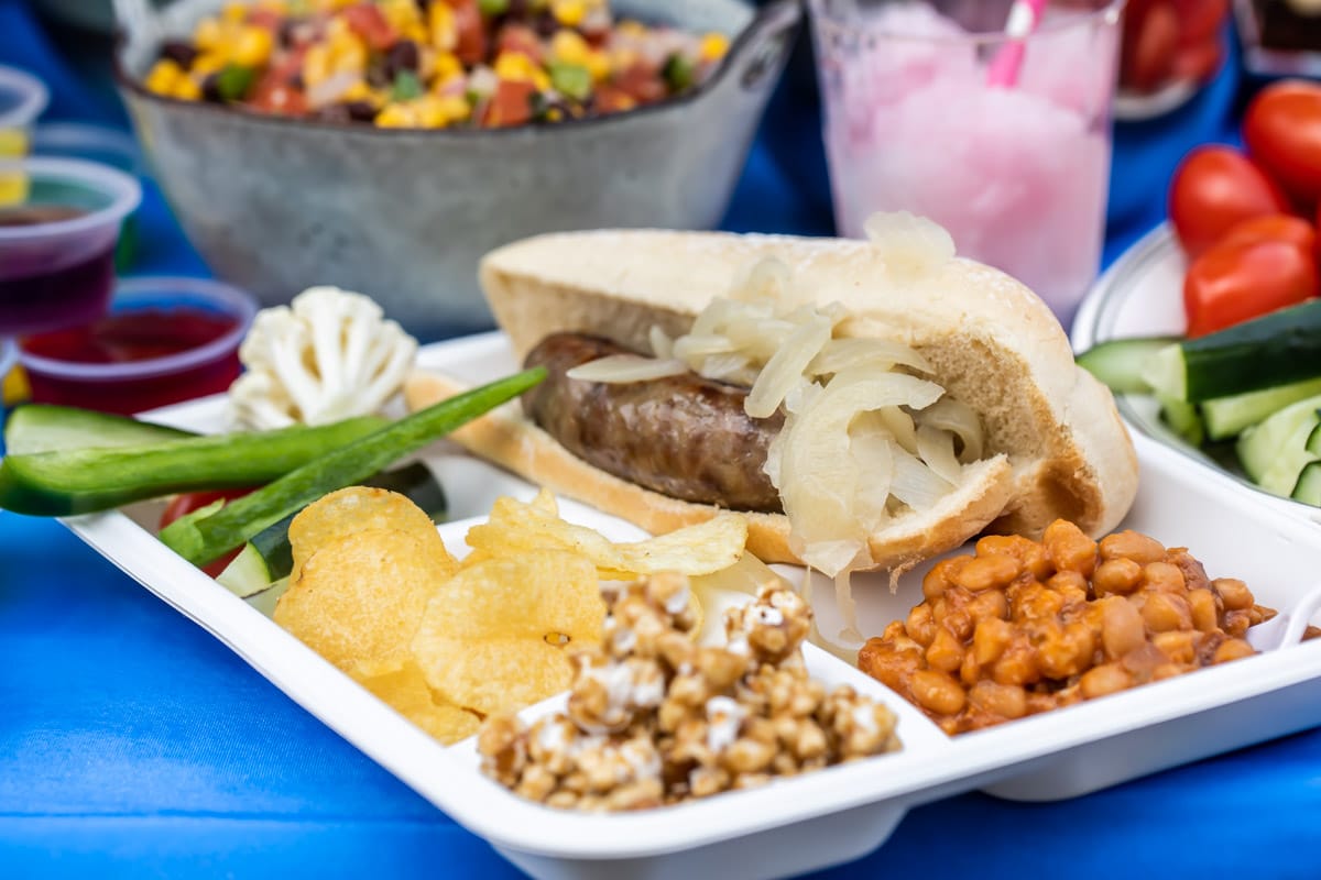 A plate at a tailgate with a beer brat, vegetables, caramel corn, chips and baked beans.