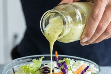 Coleslaw dressing being poured onto a large clear bowl of cabbage.