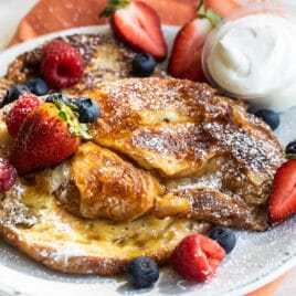 Croissant French toast on a white plate with fruit and whipped cream.