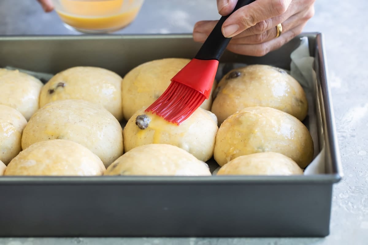 Butter being spread on uncooked hot cross buns in a silver baking pan.
