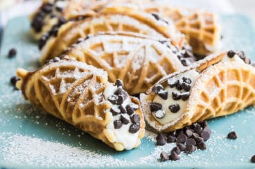 Mock italian cannoli with pizzelle on top of a blue cutting board covered in powdered sugar.