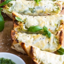 Slices of pesto cheese bread surrounded by basil leaves on a wooden cutting board.
