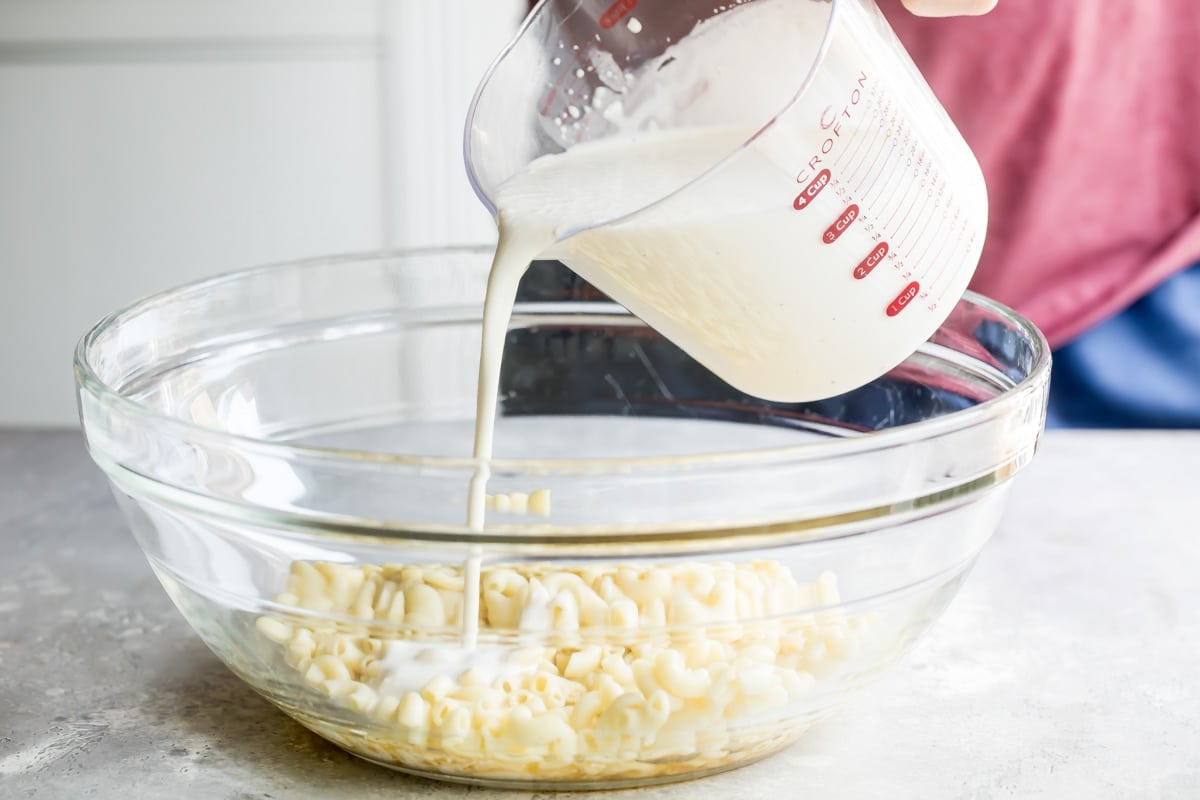 Cream being poured onto cooked macaroni pasta in a clear bowl.