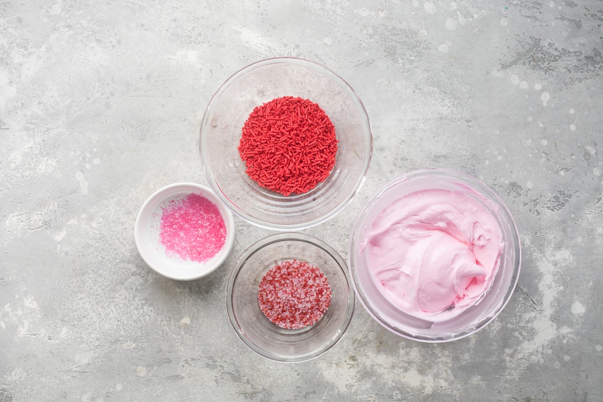 A bowl of pink royal icing next to bowls of sprinkles.