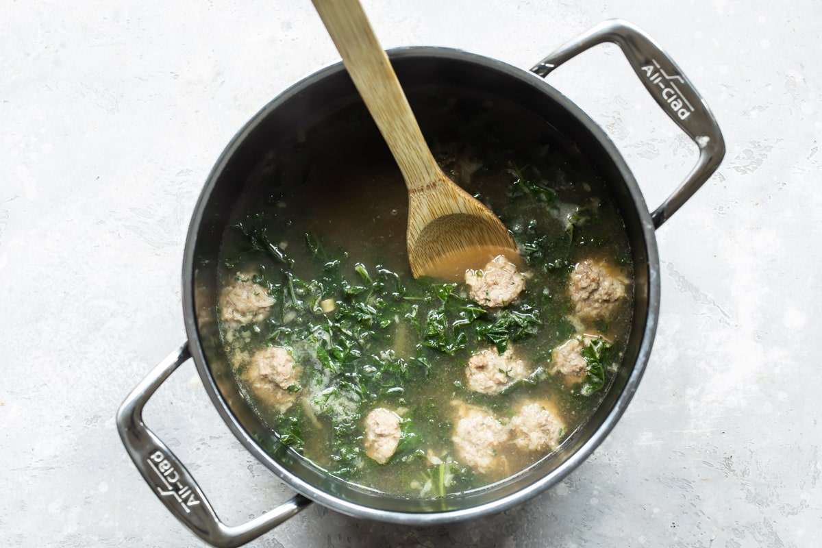 Italian wedding soup in a pot with a wooden spoon resting in it.
