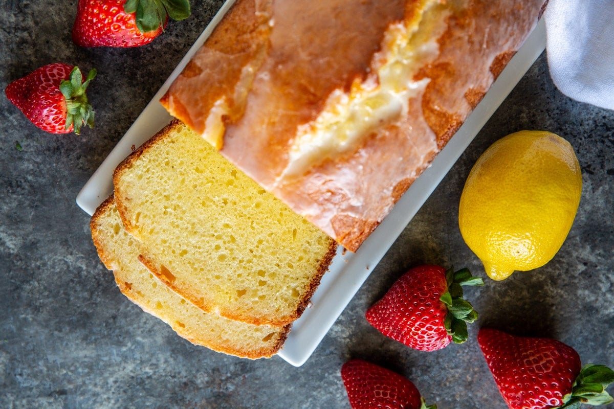Lemon Yogurt Cake with some slices cut off the loaf, surrounded by fresh strawberries and a lemon.