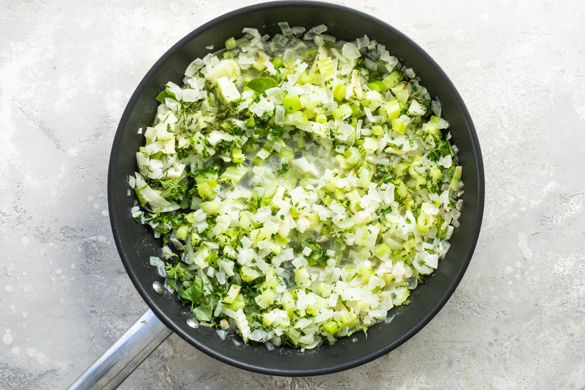 Onions and celery with fresh herbs cooking in a skillet.