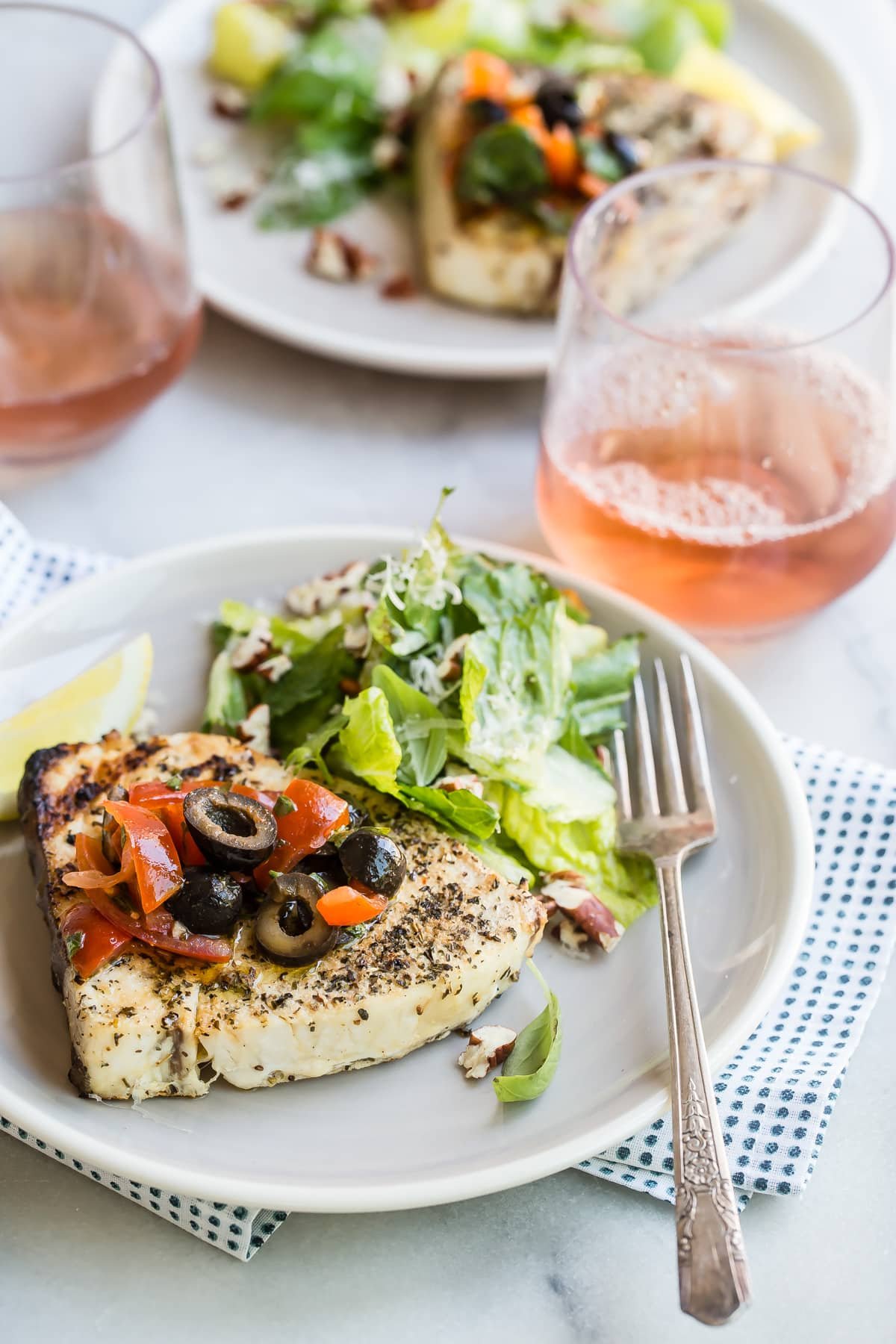 A piece of grilled swordfish on a plate with olive topping and green salad.
