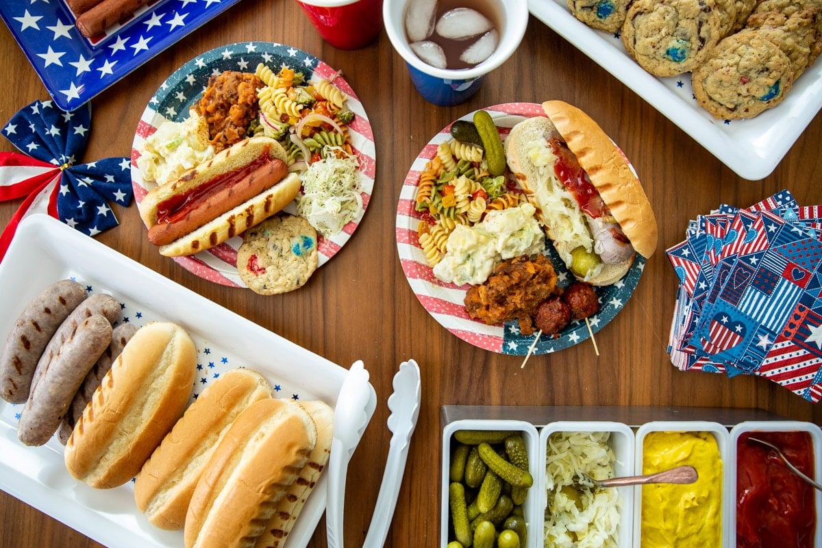 A table of 4th of July Cookout food such as brats, hot dogs, pasta salad, cookies, and drinks.