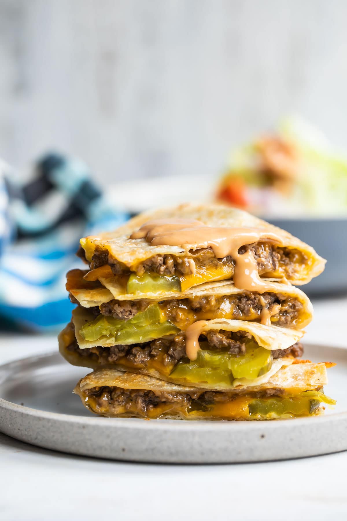 A cheeseburger quesadilla stacked on a plate with a wedge salad in the background.
