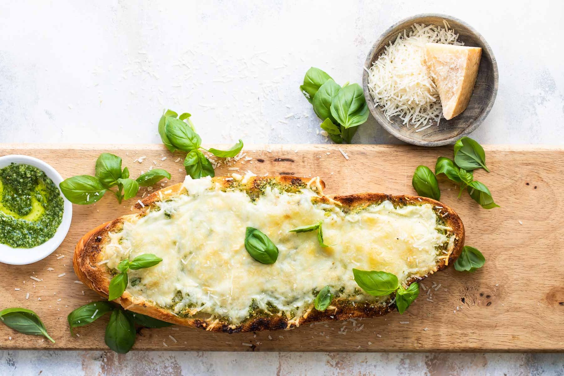 Slices of pesto cheese bread surrounded by basil leaves on a wooden cutting board.