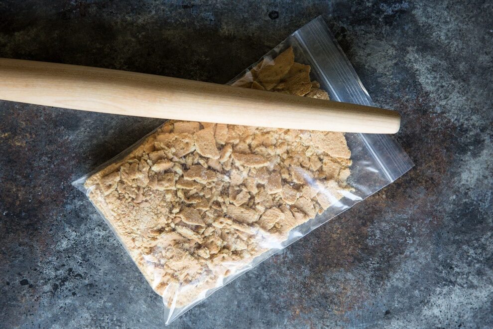 Crushing graham crackers into crumbs in a plastic bag with a rolling pin.