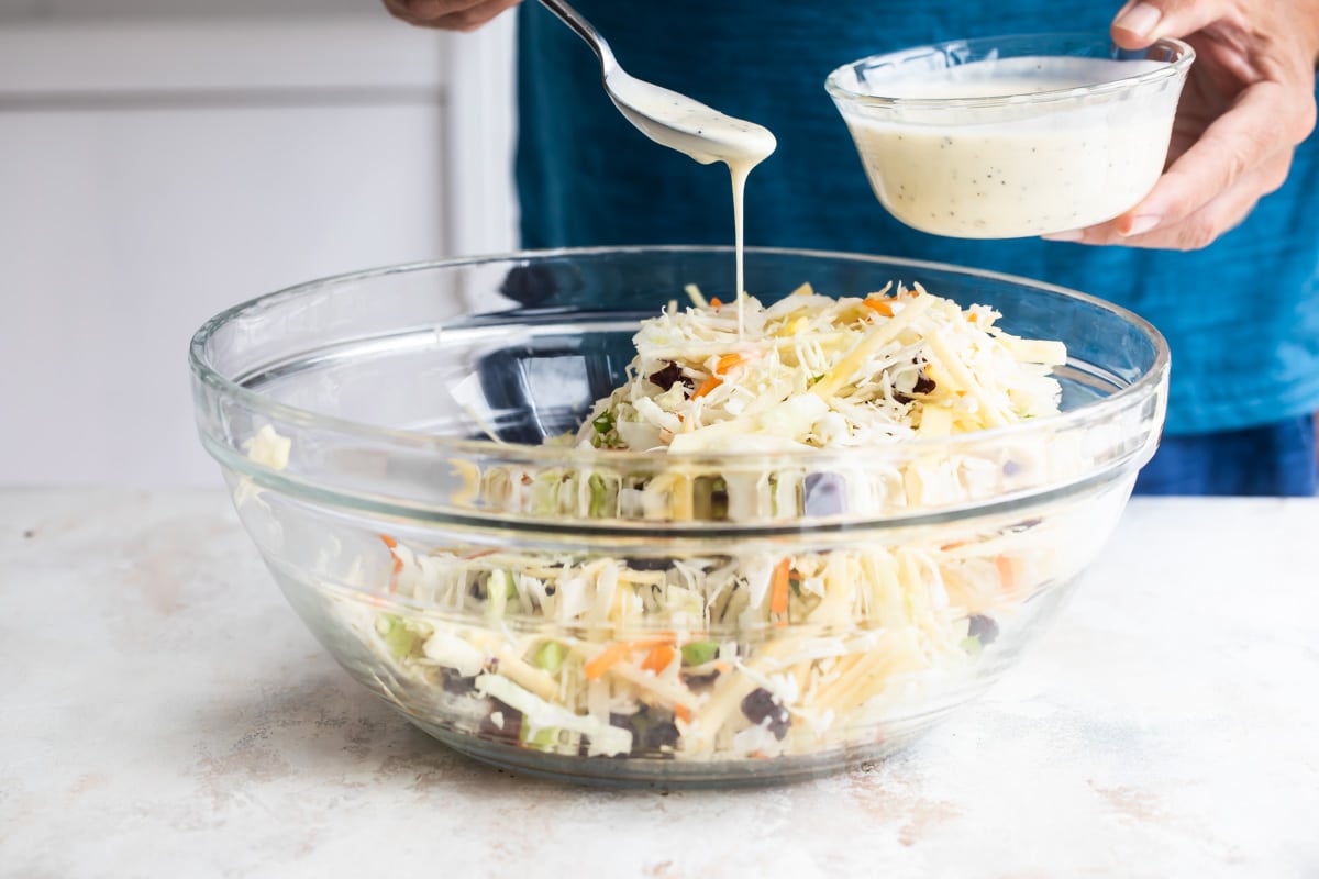 Someone drizzling dressing over apple coleslaw in a clear bowl.