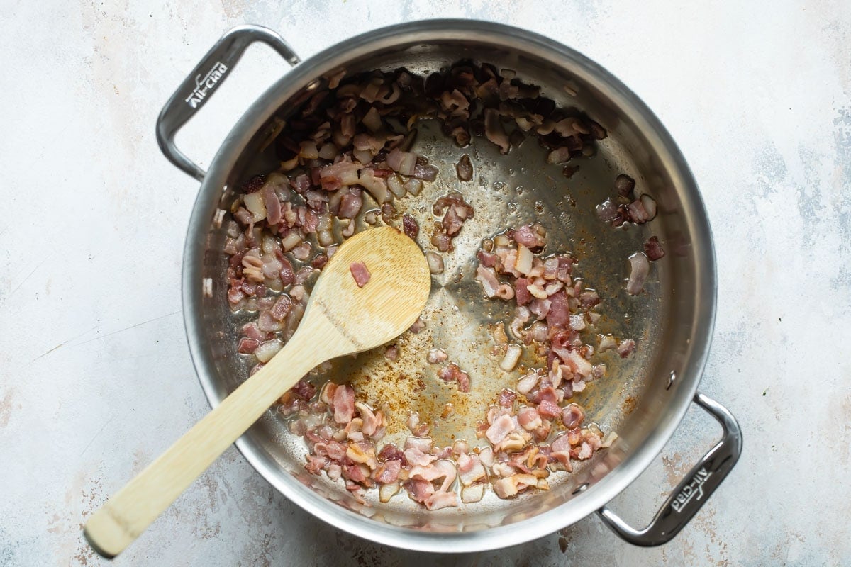 Bacon pieces being fried in a pot with a wooden spoon resting in it.