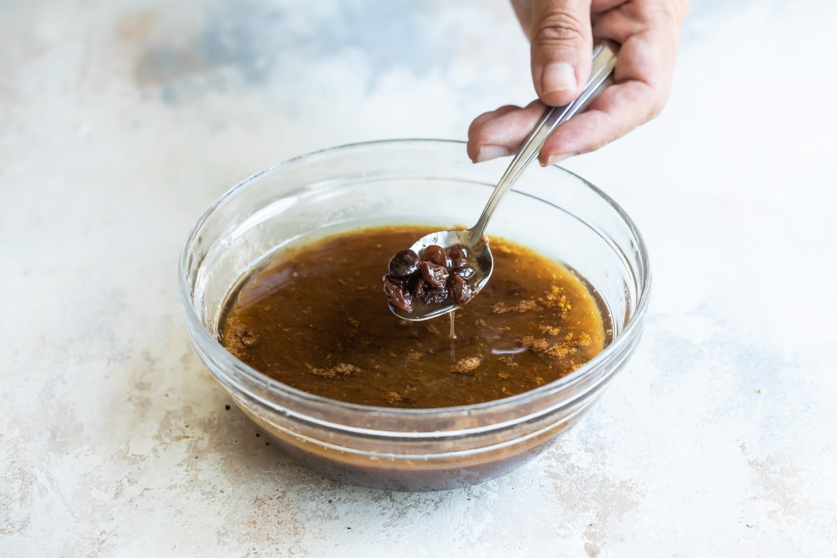 Someone holding a spoonful of soaked raisins over a clear bowl filled with brown liquid.