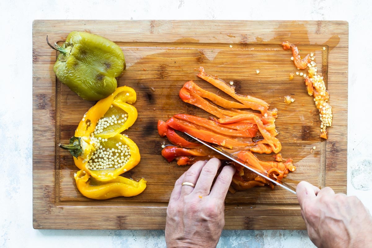 Someone slicing red, yellow, and green roasted peppers on a wood cutting board.