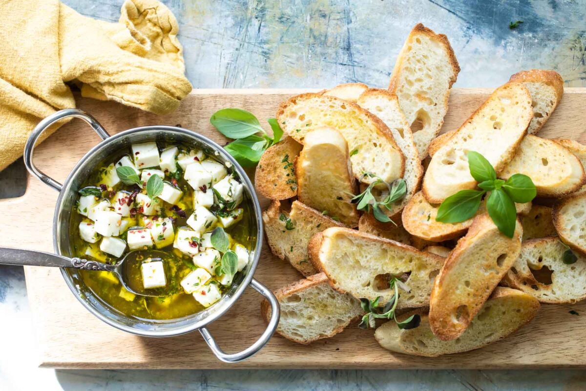 Marinated feta in a silver bowl on a wood board with slices of bread.