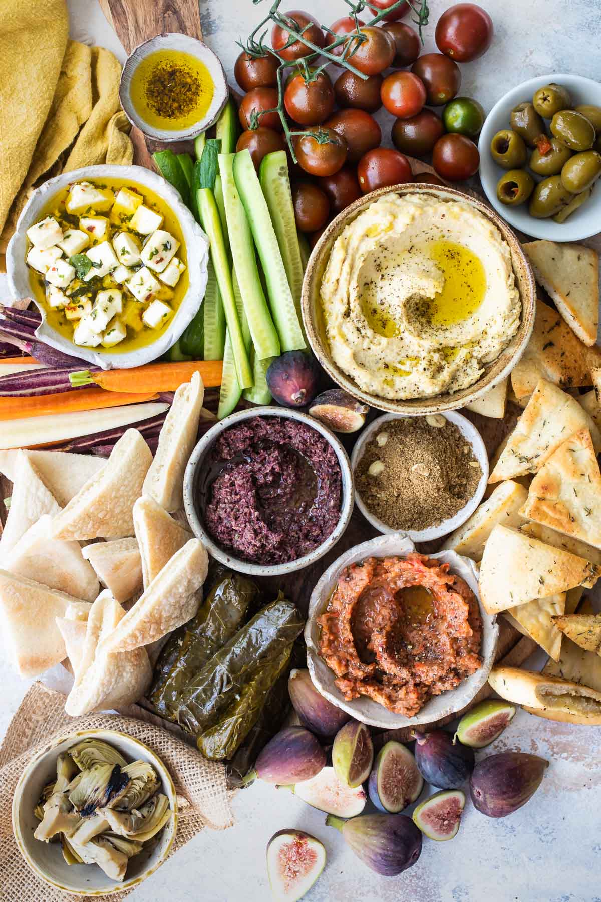 Dukkah, eggplant dip, hummus, artichokes, and olives in bowls with tomatoes, pita chips, pita bread, cucumbers and carrot sticks.