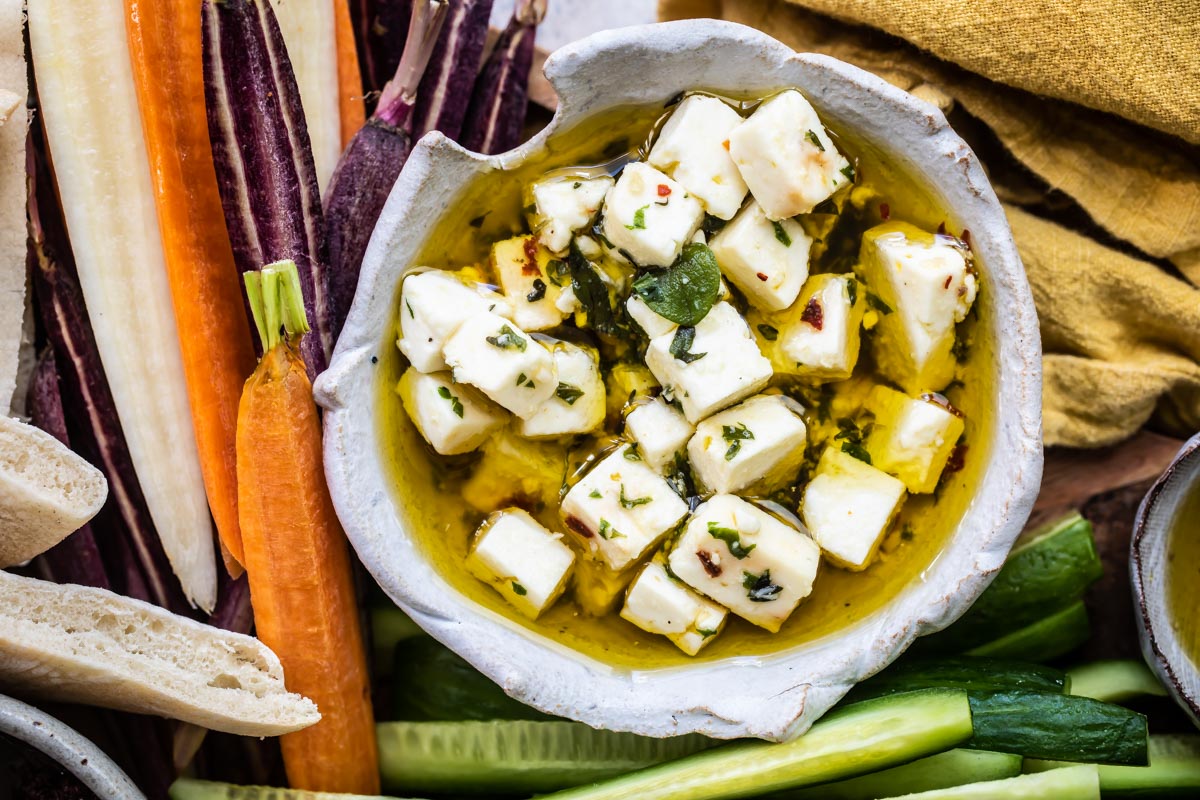 Marinated feta in a bowl surrounded by sliced carrots and cucumbers.