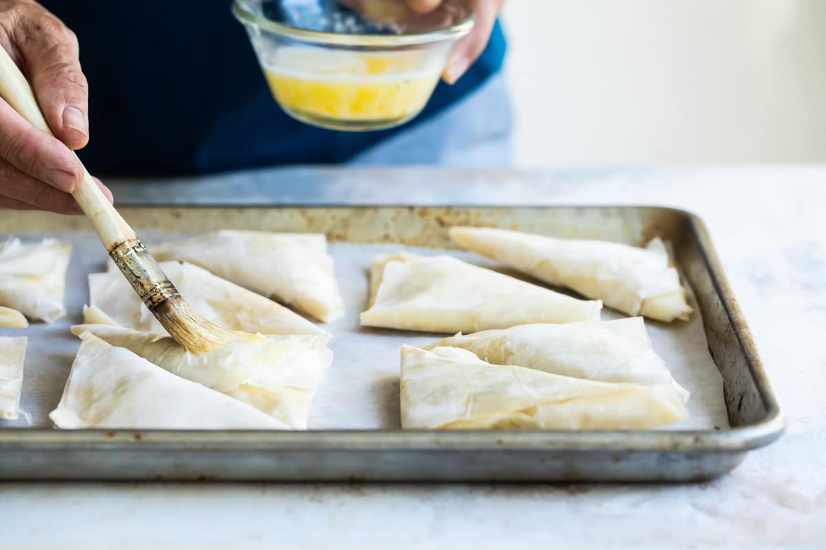 Someone brushing melted butter onto prebaked three cheese spanakopita triangles on a baking sheet.