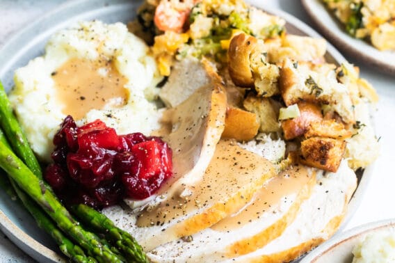 A plate of roasted turkey with gravy, mashed potatoes, bread stuffing, veggie casserole, cranberry sauce with apples, and roasted asparagus.