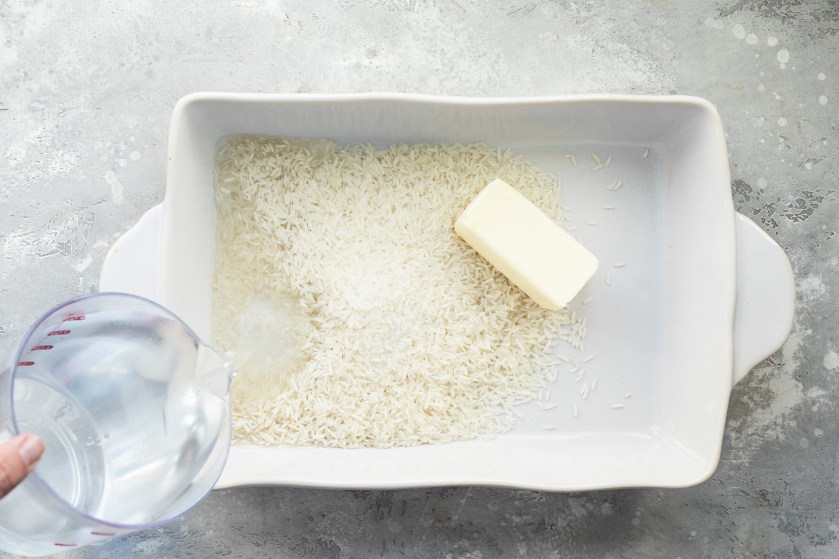Grains of rice and butter in a white baking dish with water being poured into it.